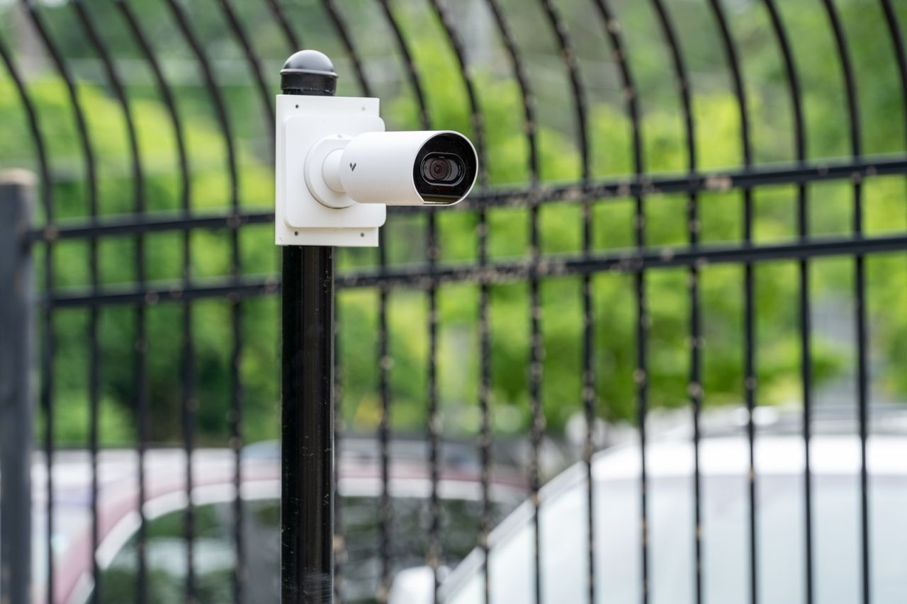 Verkada bullet security camera mounted on a black pole near a metal fence outdoors.