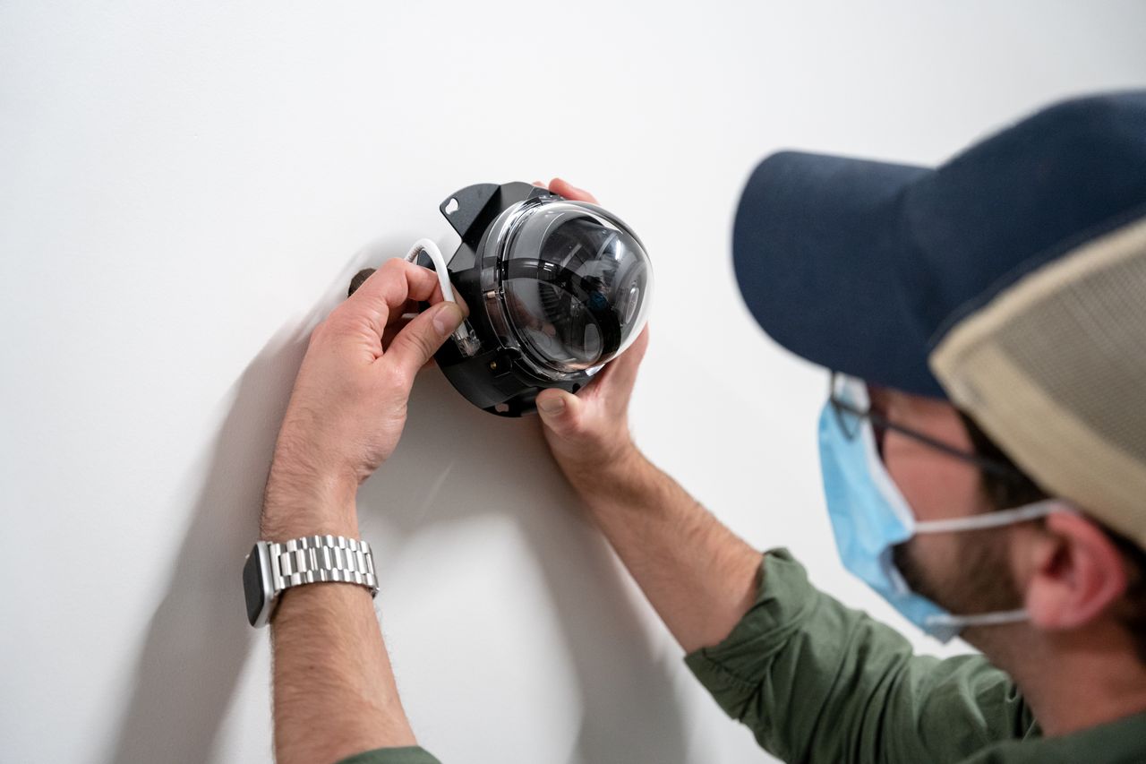 Technician wearing a face mask installs a dome security camera on a white wall.