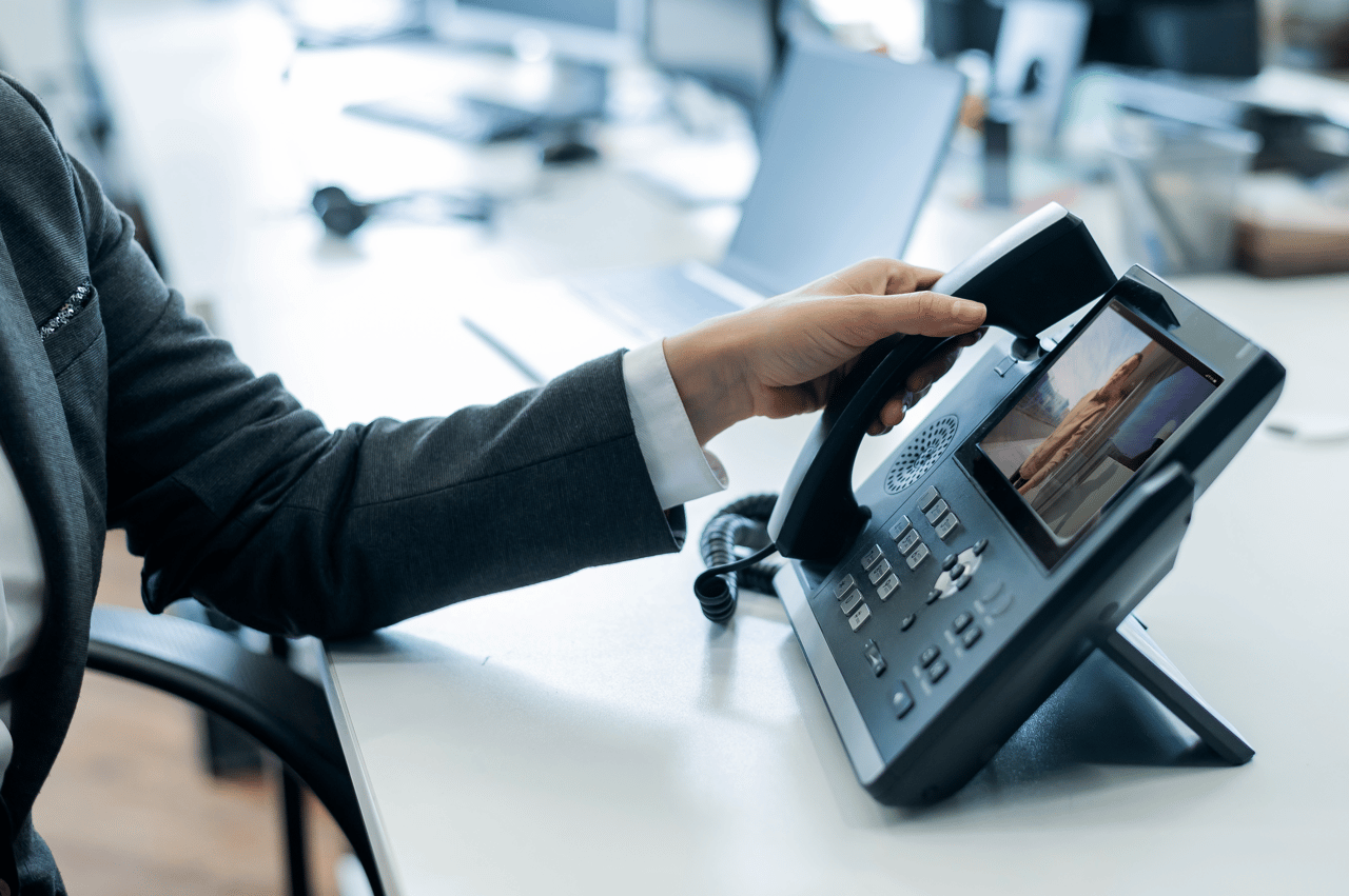 A person in a suit picks up a video desk phone in a modern office.
