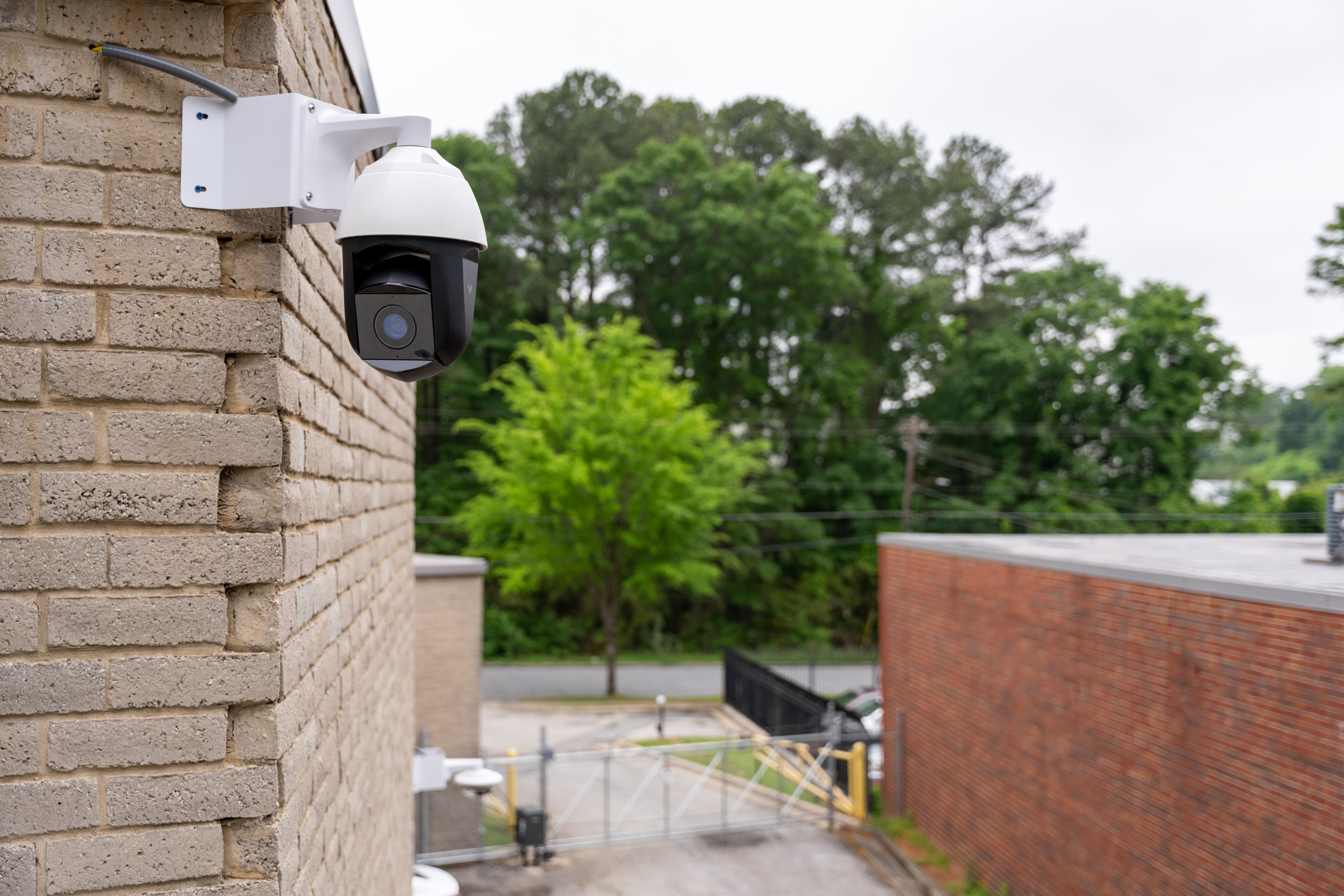 Verkada PTZ security camera mounted on a brick exterior wall overlooking a gated perimeter.